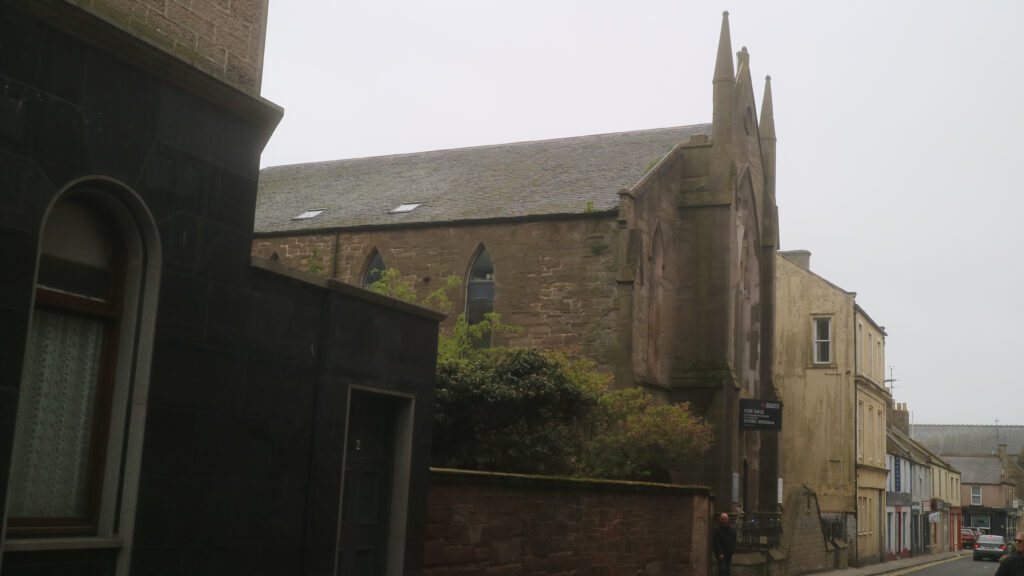 Colour photograph of street, dominated by the side and front of a red sandstone church, three small spires above the entrance silhouetted against a uniformly grey sky.