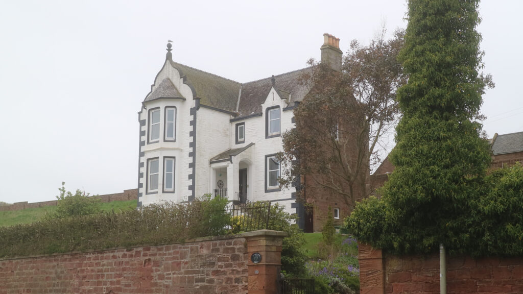 Colour photograph of large detached house, walls painted white, standing back and around 20 feet higher than the unseen road below, separated by a garden, and a red sandstone wall topped by a hedge, broken by a gateway.