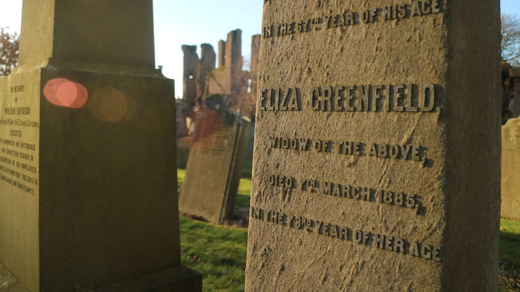 Close up of grave stone, highlighting the name 'Eliza Greenfield / Widow of the Above / Died 7th March 1885 / In the 78th year of her age'. Surrounded by other graves, and in the background the ruins of an abbey, against a blue sky. Overlapping red circles of refracted light appear against the side of the neighbouring grave stone that is in shadow.