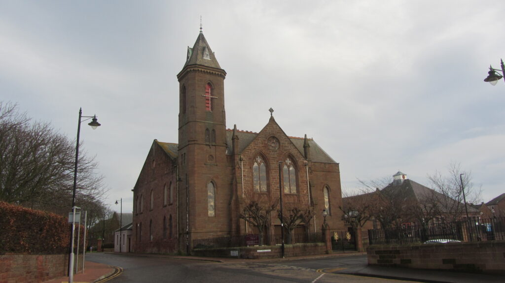 Colour photograph of red standstone church, with spire, standing at crossroads. Smaller building visible behind leafless tree to the right. Hedge and leafless trees on the left, old-fashioned street lights silhouetted against the grey sky.