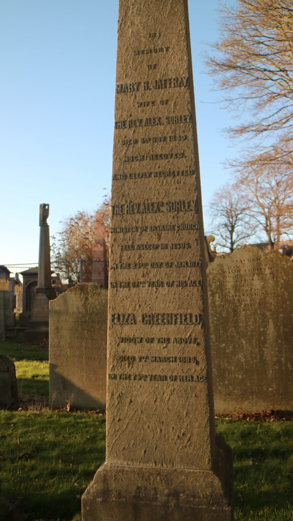 Colour photograph of tall gravestone, bearing names and dates, surrounded by other graves, against a clear blue sky, and branches of trees suggesting autumn.