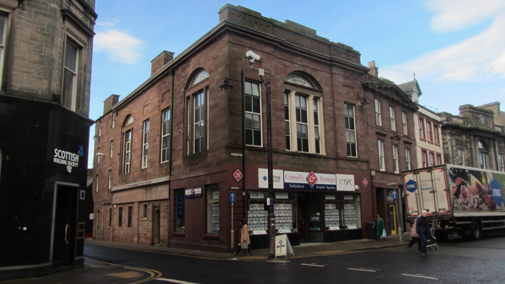 Colour photo of 19th-century red sandstone building at a town crossroads. Ground floor appears to be occupied by estate agents' shop. Upper floors have large windows, suggesting former use as a public hall.