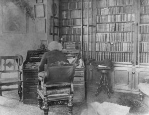 Black and white photograph, Douglass, with his back to the camera, sitting at his desk in his library; a violin is visible on his desk.