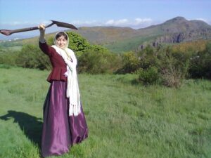 Woman in 19th-century dress brandishing a spade, with Arthur's Seat and Salisbury Crags in the background.